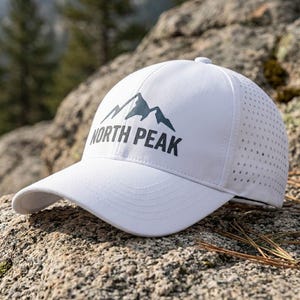 May include: A white baseball cap featuring a dark gray mountain graphic and the text NORTH PEAK printed on the front. The cap has a curved brim and perforated side panels for ventilation, constructed with a structured crown and stitched detailing.