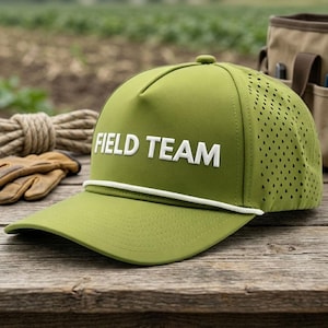 May include: An olive green baseball cap featuring the white text FIELD TEAM on the front panel. The cap includes a white rope detail across the base of the crown and perforated side panels for ventilation, constructed with a structured front.