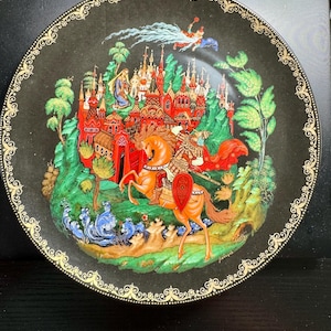 May include: A round black decorative plate featuring a detailed illustration of a knight on a brown horse in front of a red castle. The scene includes green trees, blue water, and a figure flying above. The rim has a gold filigree pattern.