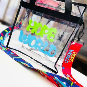 May include: Clear tote bag with black trim and handles. The bag features the words "HOPE WORLD" in green and blue. A red lanyard with "HOPE WORLD" printed on it is attached. A colorful patterned strap is also included.