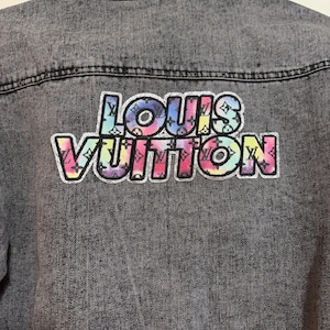 Könnte beinhalten: Graue Jeansjacke mit einem farbenfrohen "LOUIS VUITTON" Logo-Patch auf dem Rücken. Das Logo zeigt ein Regenbogen-Tie-Dye-Muster. Die Jacke hat einen klassischen Kragen und eine Knopfleiste.