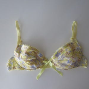 May include: A yellow underwire bra featuring a delicate floral pattern in shades of purple and light brown. The garment includes adjustable yellow shoulder straps and a small yellow satin ribbon bow centered at the bridge between the cups.
