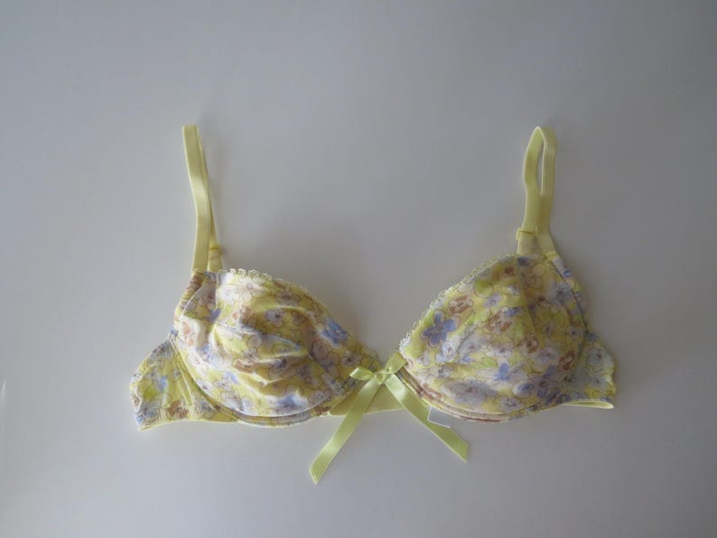 May include: A yellow underwire bra featuring a delicate floral pattern in shades of purple and light brown. The garment includes adjustable yellow shoulder straps and a small yellow satin ribbon bow centered at the bridge between the cups.
