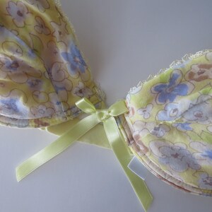 May include: A yellow underwire bra featuring a floral pattern in shades of blue, white, and brown. The garment includes a small, light green satin ribbon bow at the center gore and delicate white lace trim along the top edges of the cups.