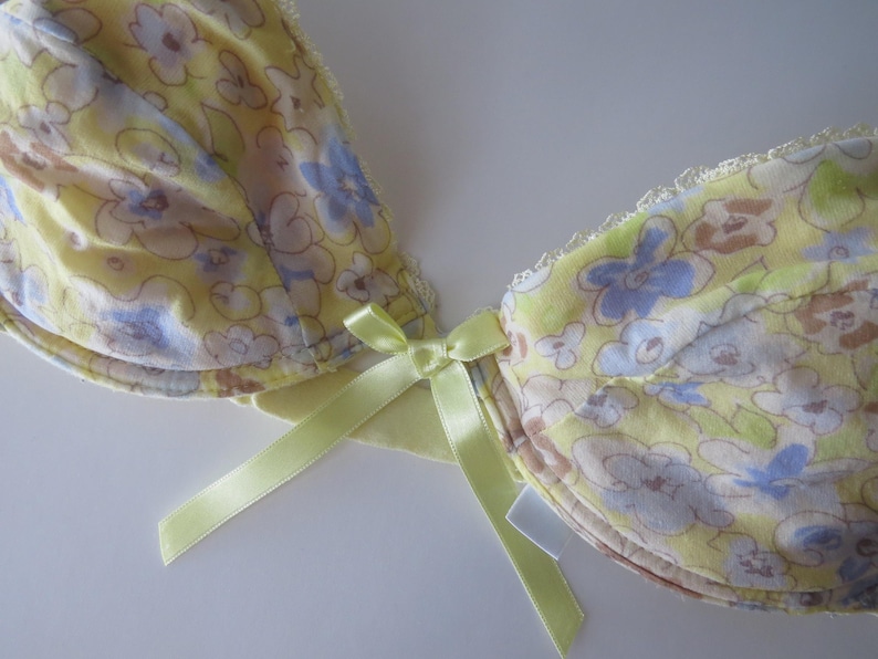May include: A yellow underwire bra featuring a floral pattern in shades of blue, white, and brown. The garment includes a small, light green satin ribbon bow at the center gore and delicate white lace trim along the top edges of the cups.