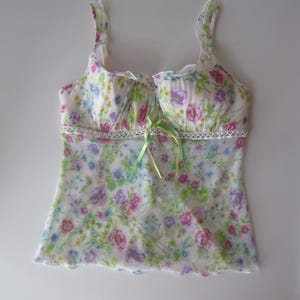 May include: A sleeveless camisole top featuring a white base with a colorful floral print in shades of pink, purple, blue, and green. The garment includes ruffled shoulder straps, a lace trim detail at the waist, and a small lime green ribbon bow.