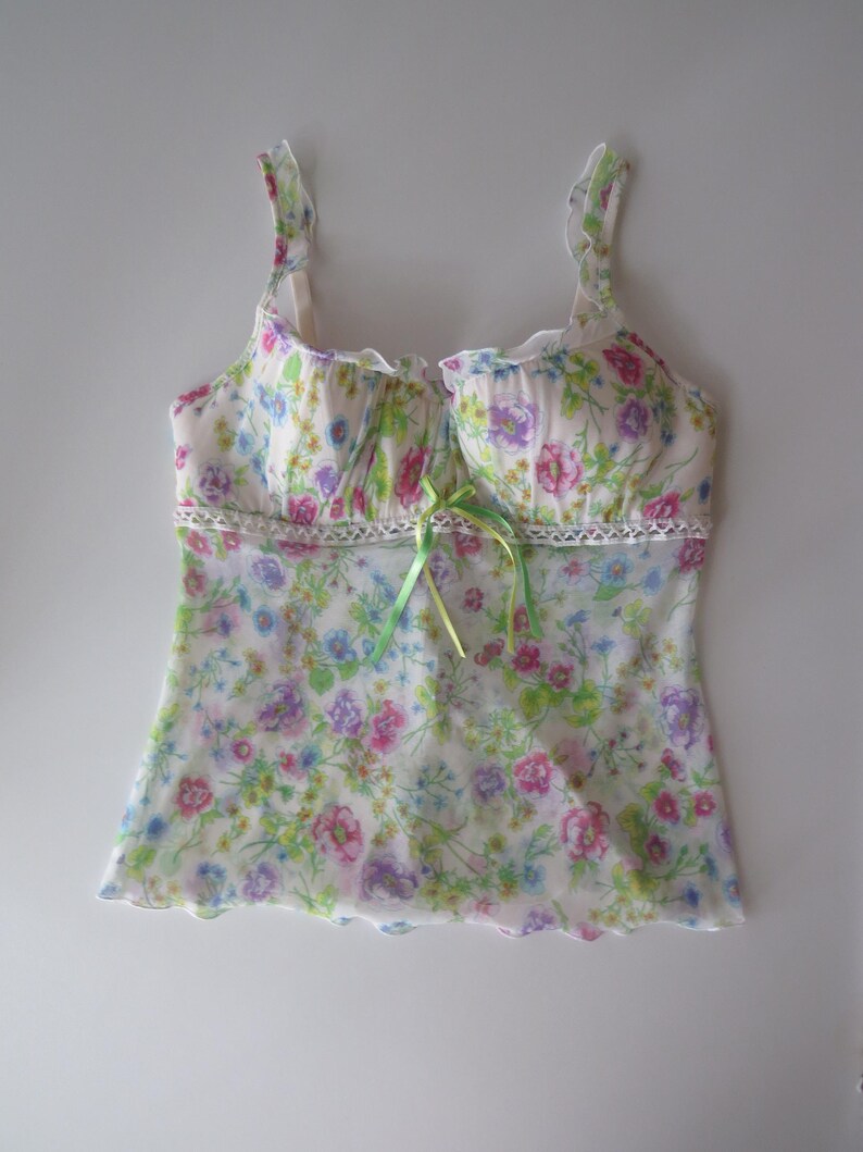 May include: A sleeveless camisole top featuring a white base with a colorful floral print in shades of pink, purple, blue, and green. The garment includes ruffled shoulder straps, a lace trim detail at the waist, and a small lime green ribbon bow.