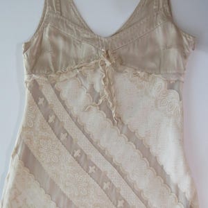 May include: A sleeveless beige dress featuring a V-neckline with a small central tie. The bodice is made of solid fabric, while the lower section is composed of diagonal panels of sheer fabric alternating with intricate cream-colored lace trim.