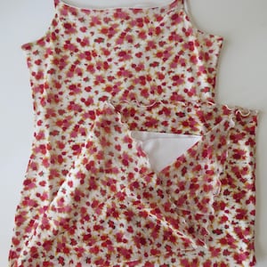 May include: A sleeveless camisole top featuring a white base with a dense floral pattern in shades of red, pink, and yellow. The garment has thin spaghetti straps, a straight neckline, and a ruffled lettuce-edge hemline along the bottom.