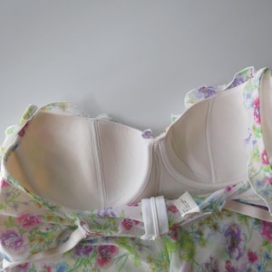 May include: A strapless bodice garment featuring a white base with a colorful floral print in shades of pink, purple, blue, and green. The interior is lined with beige fabric and includes molded cups with underwire support and a white back zipper.