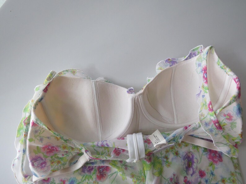 May include: A strapless bodice garment featuring a white base with a colorful floral print in shades of pink, purple, blue, and green. The interior is lined with beige fabric and includes molded cups with underwire support and a white back zipper.