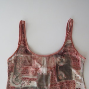May include: A sleeveless tank top featuring a scoop neckline and thin shoulder straps. The garment is made of a semi-sheer fabric with a multi-colored patchwork pattern in shades of terracotta, brown, and cream, including floral and geometric motifs.