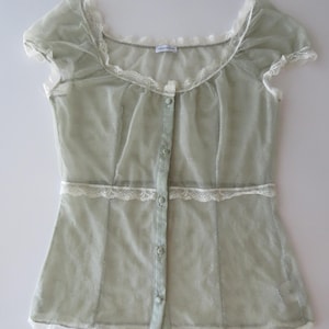 May include: A sage green sheer short-sleeve blouse featuring a scoop neckline, a front button-down closure with matching fabric buttons, and white lace trim detailing along the neckline, waist, and hem. The fabric has a subtle dotted texture.