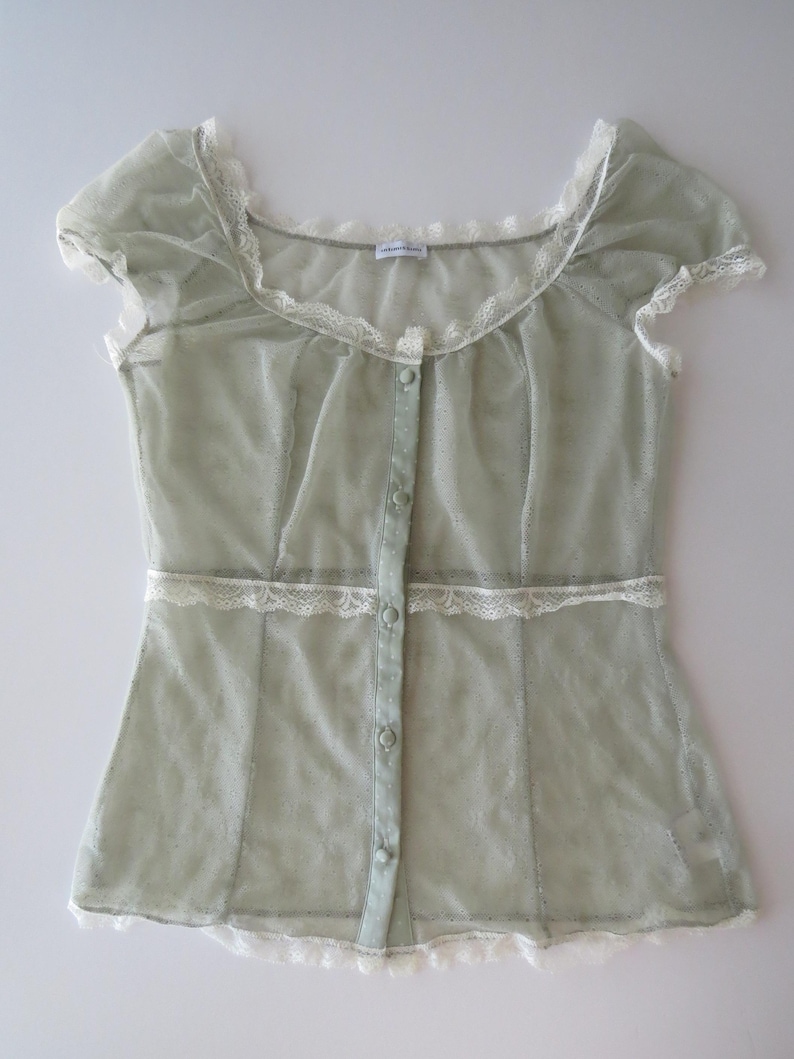 May include: A sage green sheer short-sleeve blouse featuring a scoop neckline, a front button-down closure with matching fabric buttons, and white lace trim detailing along the neckline, waist, and hem. The fabric has a subtle dotted texture.