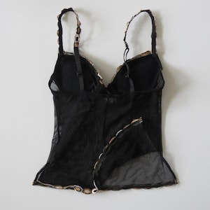 Puede incluir: Top de lencer&iacute;a negro con tirantes ajustables y un dise&ntilde;o de malla transparente. El top presenta un ribete festoneado con detalles dorados y blancos a lo largo del escote y el dobladillo. La espalda tiene un cierre de corchete y ojal.