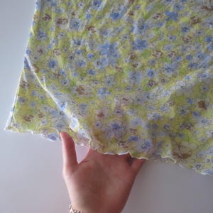 May include: A light yellow top featuring a repeating floral pattern in shades of blue, white, and brown. The garment has a lettuce-edge hemline with a wavy finish along the bottom border, showcasing a lightweight fabric with a soft, semi-sheer texture.