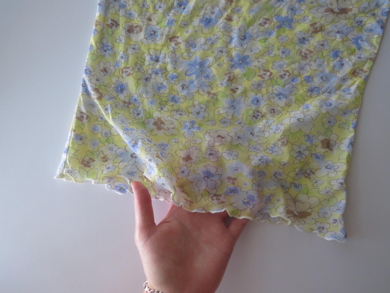 May include: A light yellow top featuring a repeating floral pattern in shades of blue, white, and brown. The garment has a lettuce-edge hemline with a wavy finish along the bottom border, showcasing a lightweight fabric with a soft, semi-sheer texture.
