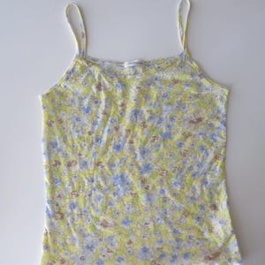 May include: A sleeveless camisole top featuring a light yellow base color with a repeating floral pattern in shades of blue, white, and brown. The garment has thin shoulder straps, a scoop neckline, and a lettuce-edge hemline at the bottom.
