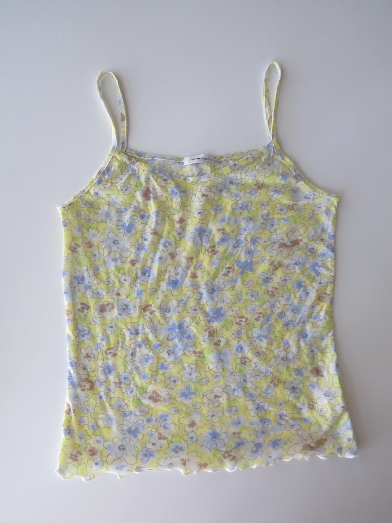 May include: A sleeveless camisole top featuring a light yellow base color with a repeating floral pattern in shades of blue, white, and brown. The garment has thin shoulder straps, a scoop neckline, and a lettuce-edge hemline at the bottom.