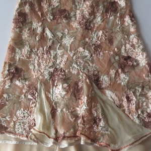 May include: A knee-length skirt featuring a sheer, tan-colored overlay with an intricate, raised floral embroidery in shades of cream, copper, and brown. The skirt has a layered design with a solid, smooth cream-colored lining visible at the hem.