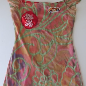 May include: A sleeveless dress featuring a multi-colored tie-dye pattern in shades of orange, pink, and purple, overlaid with a light green floral line design. The garment includes a circular red patch with white floral embroidery and a small car patch.