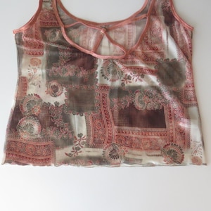 May include: A sleeveless tank top featuring a patchwork-style print in shades of terracotta, brown, and cream. The garment has a scoop neckline with a thin strap detail across the upper chest and a lettuce-edge hemline along the bottom.