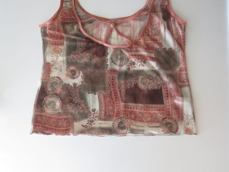 May include: A sleeveless tank top featuring a patchwork-style print in shades of terracotta, brown, and cream. The garment has a scoop neckline with a thin strap detail across the upper chest and a lettuce-edge hemline along the bottom.