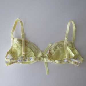 May include: A pale yellow bra featuring adjustable shoulder straps and a central ribbon bow detail. The band and edges are decorated with a delicate floral pattern in shades of purple, blue, and white, with a hook-and-eye closure at the back.