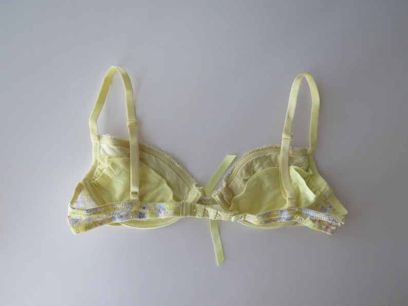 May include: A pale yellow bra featuring adjustable shoulder straps and a central ribbon bow detail. The band and edges are decorated with a delicate floral pattern in shades of purple, blue, and white, with a hook-and-eye closure at the back.