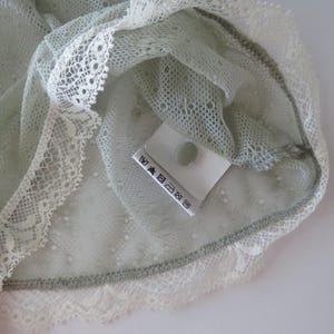 May include: A sage green lace garment featuring a delicate, semi-sheer mesh fabric with a repeating dotted pattern. The edges are finished with a wide, scalloped white lace trim. A white care label with black laundry symbols is attached inside.