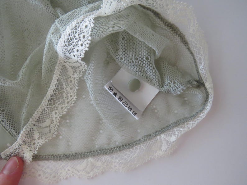 May include: A sage green lace garment featuring a delicate, semi-sheer mesh fabric with a repeating dotted pattern. The edges are finished with a wide, scalloped white lace trim. A white care label with black laundry symbols is attached inside.