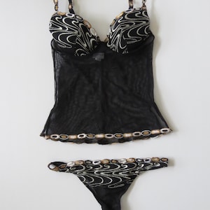 Puede incluir: Conjunto de lencer&iacute;a negra con top transparente y tanga a juego. El conjunto est&aacute; adornado con intrincados bordados en tonos dorados y plateados, creando un patr&oacute;n decorativo. Las correas y los bordes est&aacute;n adornados con detalles similares.