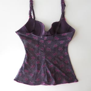 May include: A dark purple camisole with a built-in underwire bra featuring adjustable straps with ruffled edges. The garment is made of a sheer, dark purple mesh fabric adorned with a repeating pattern of small, vibrant pink floral clusters.