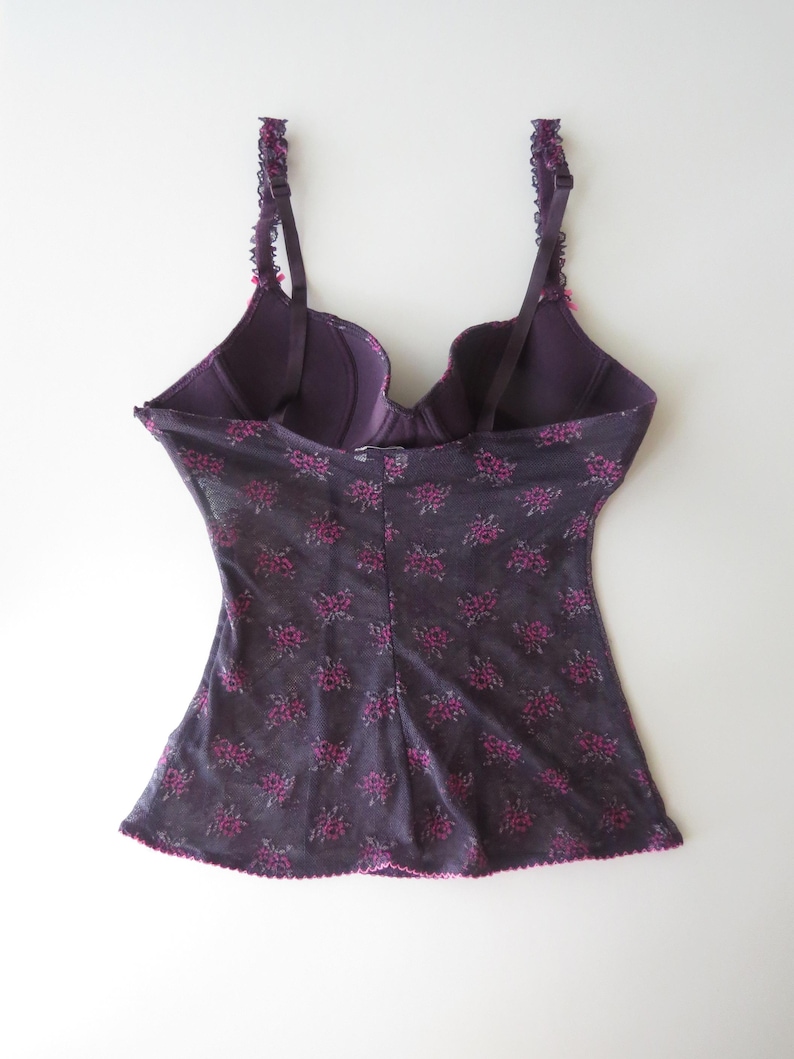May include: A dark purple camisole with a built-in underwire bra featuring adjustable straps with ruffled edges. The garment is made of a sheer, dark purple mesh fabric adorned with a repeating pattern of small, vibrant pink floral clusters.