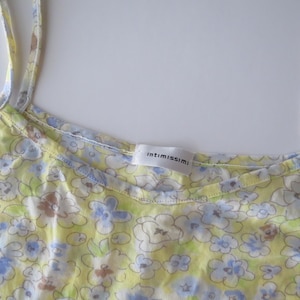 May include: A yellow camisole with thin shoulder straps featuring a light blue, white, and brown floral pattern. The garment has a scoop neckline and a white interior label with the brand name intimissimi printed in black text at the center.
