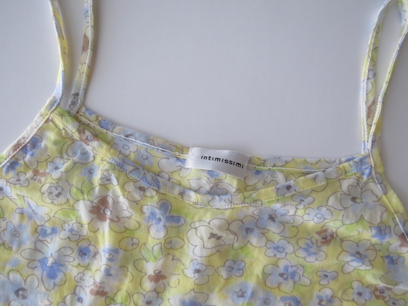 May include: A yellow camisole with thin shoulder straps featuring a light blue, white, and brown floral pattern. The garment has a scoop neckline and a white interior label with the brand name intimissimi printed in black text at the center.