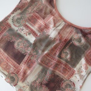 May include: A sleeveless top featuring a sheer, lightweight fabric with a multi-colored patchwork pattern in shades of terracotta, brown, and cream. The garment has a round neckline with a solid terracotta-colored trim along the collar and armholes.