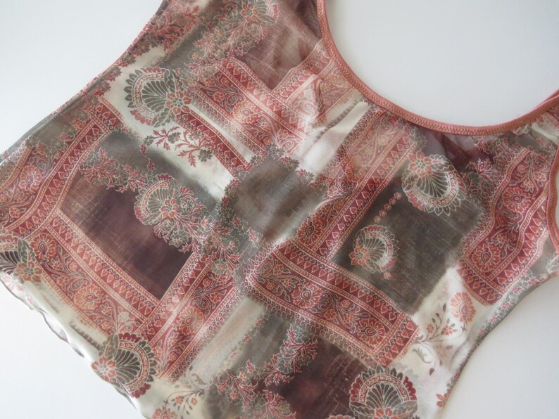 May include: A sleeveless top featuring a sheer, lightweight fabric with a multi-colored patchwork pattern in shades of terracotta, brown, and cream. The garment has a round neckline with a solid terracotta-colored trim along the collar and armholes.