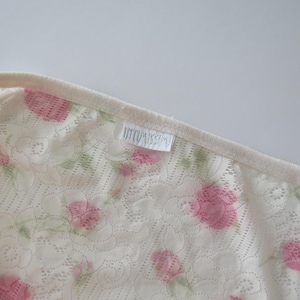 Pu&ograve; includere: Una canotta in pizzo color crema con un delicato motivo floreale di rose rosa e foglie verdi. Il capo presenta sottili spalline regolabili e un'etichetta in tessuto bianco con la scritta Intimissimi cucita nello scollo.