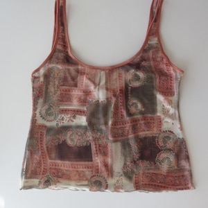 May include: A sleeveless tank top featuring a sheer, lightweight fabric with a multi-colored patchwork print in shades of terracotta, brown, and cream. The garment has a scoop neckline, wide shoulder straps, and a straight hemline with a subtle wavy edge.