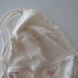 Pu&ograve; includere: Un bralette triangolare bianco con sottili doppie spalline e un delicato rivestimento in pizzo con un sottile motivo floreale rosa. Il capo presenta un bordo smerlato lungo lo scollo e una struttura in tessuto morbido e semi-trasparente.