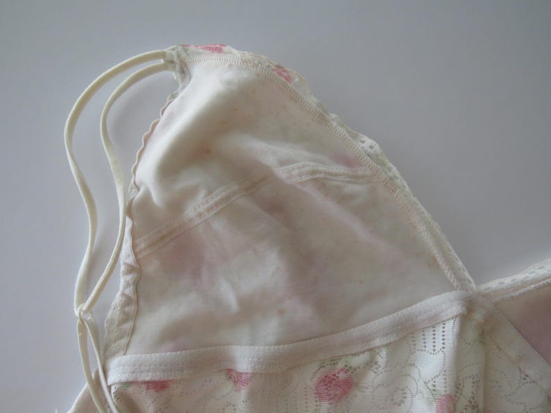 Pu&ograve; includere: Un bralette triangolare bianco con sottili doppie spalline e un delicato rivestimento in pizzo con un sottile motivo floreale rosa. Il capo presenta un bordo smerlato lungo lo scollo e una struttura in tessuto morbido e semi-trasparente.
