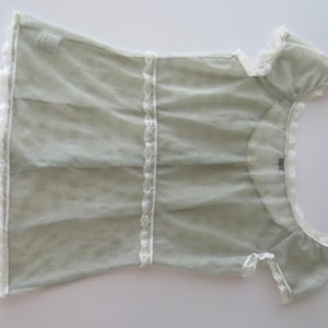 May include: A sage green short-sleeved top featuring a sheer, textured fabric with white lace trim along the neckline, sleeves, and hem. The garment includes a vertical white lace panel down the center and a small tag at the inner neckline.