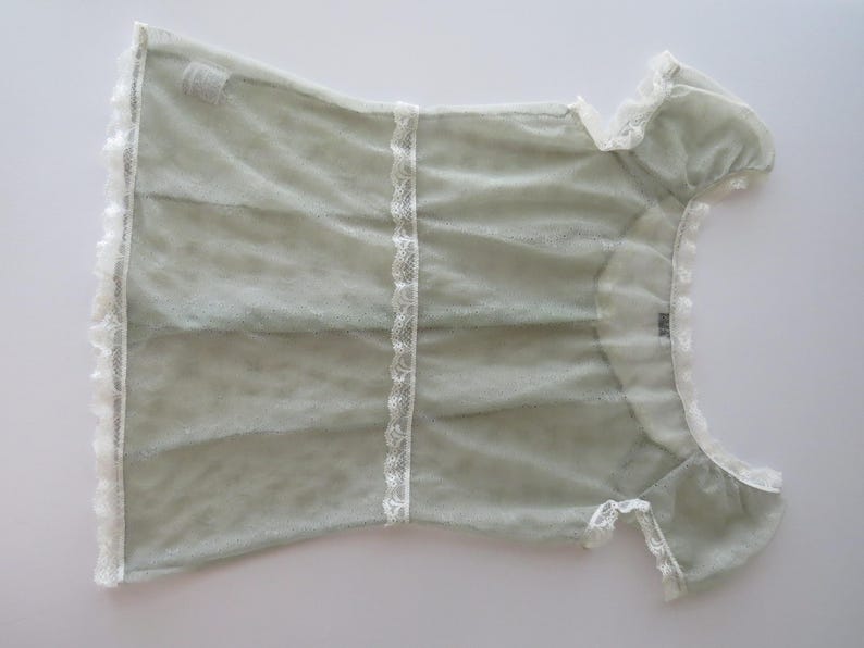 May include: A sage green short-sleeved top featuring a sheer, textured fabric with white lace trim along the neckline, sleeves, and hem. The garment includes a vertical white lace panel down the center and a small tag at the inner neckline.