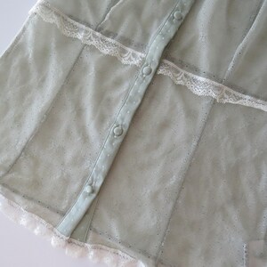 May include: A sage green button-down shirt featuring a sheer, textured fabric with a subtle dotted pattern. The garment is accented with horizontal white lace trim across the chest and a matching white lace border along the bottom hem.