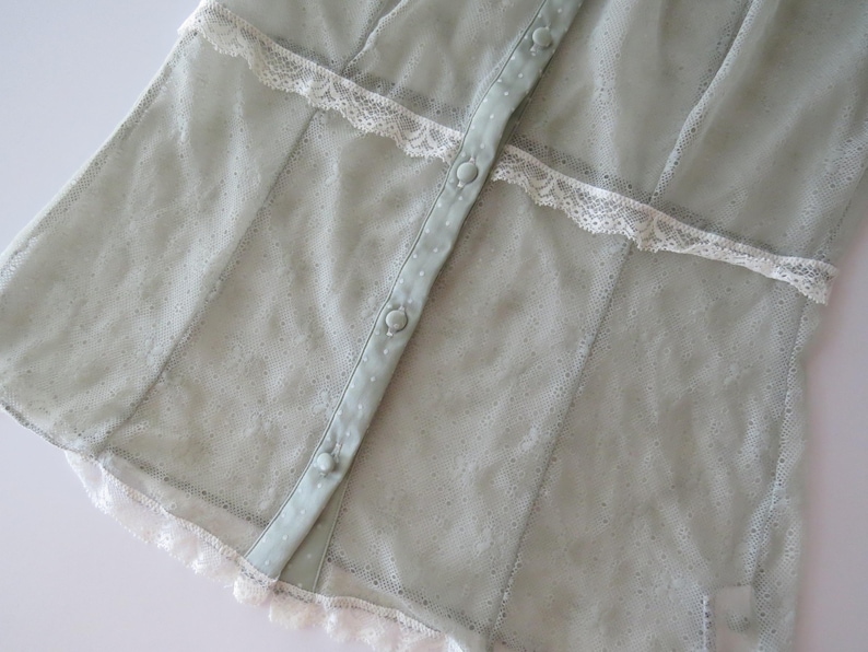 May include: A sage green button-down shirt featuring a sheer, textured fabric with a subtle dotted pattern. The garment is accented with horizontal white lace trim across the chest and a matching white lace border along the bottom hem.