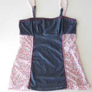 May include: A sleeveless tank top featuring a solid dark gray center panel and side panels with a white and red floral print. The garment includes thin spaghetti straps in the same floral pattern and red decorative stitching along the neckline.