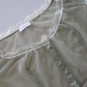 May include: A sage green sheer mesh top featuring a delicate cream-colored lace trim along the neckline and shoulders. The garment includes a central placket with matching sage green buttons and a small white tag labeled Intimissimi.