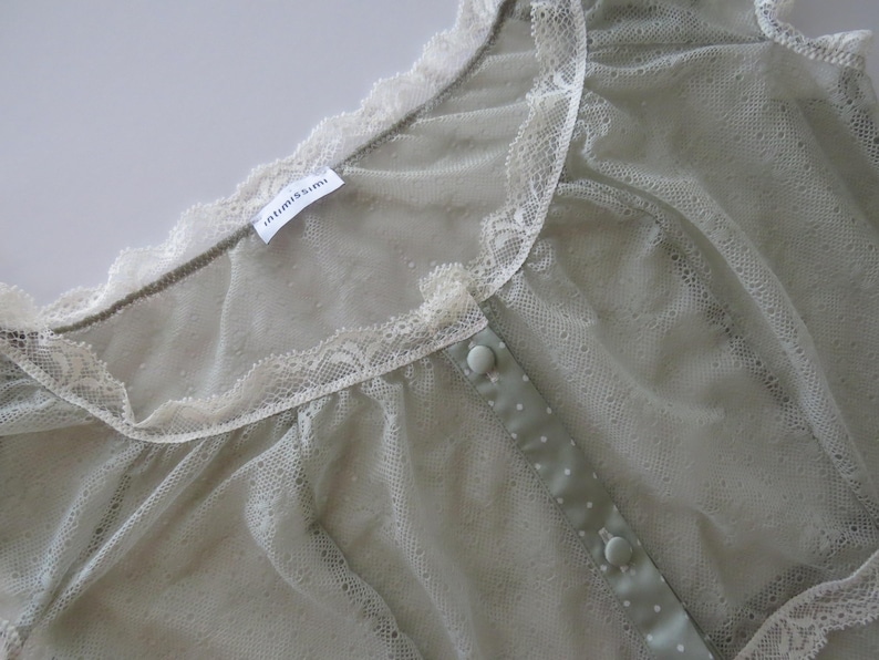 May include: A sage green sheer mesh top featuring a delicate cream-colored lace trim along the neckline and shoulders. The garment includes a central placket with matching sage green buttons and a small white tag labeled Intimissimi.