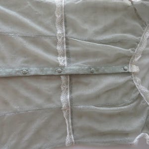 May include: A light sage green sheer mesh blouse featuring a delicate white lace trim along the vertical seams. The garment includes a central button placket with matching sage green fabric-covered buttons and a soft, semi-transparent texture.